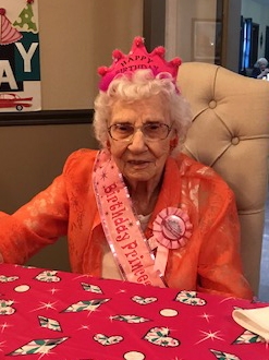Bertha sits at a table with a pink table cloth. She wears a pink crown, ribbon, and sash all referencing her birthday.