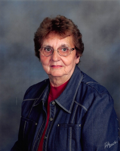 Deloris sits in front of a blue-gray background, wearing a red blouse, denim jacket, and glasses.