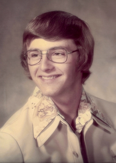An old photo that looks to be from the 1970's. A young David smiles broadly, looking to the left. He wears collared shirts and wire-framed glasses.
