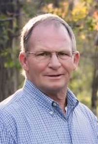 Robert smiles at the camera, wearing a blue plaid button-up in front of autumnal trees.