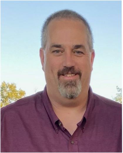 Mark smiles to the camera. The sky is behind him and he wears a red polo.