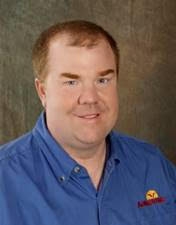 Jeff poses for a photo in front of a brown background. He wears a blue polo.