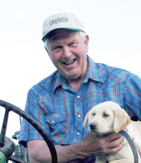 Howard sits at a tractor wheel, beaming and holding a puppy.