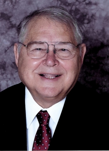 John smiles widely in front of a mottled background. He wears wide, wire-rimmed glasses and a suit and tie.