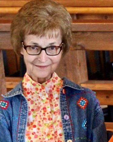 Marilyn smiles at the camera. She wears rectangular framed glasses, an orange blouse, and embroidered denim jacket.