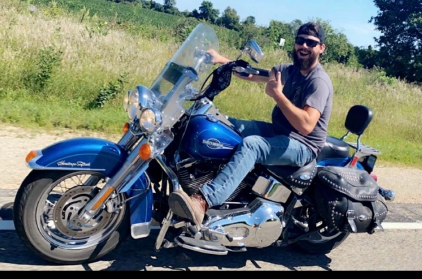 John rides atop his blue motorcycle, giving a large smile and thumbs up.